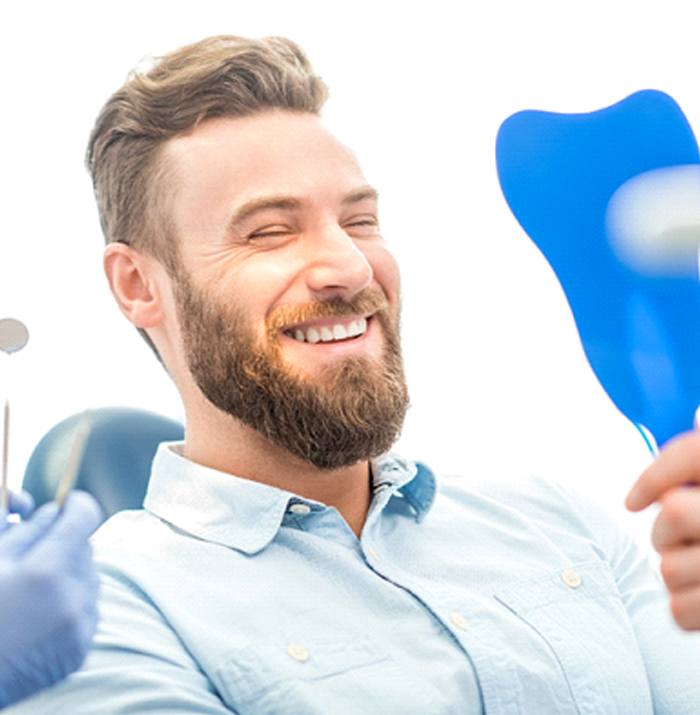 A young male with a beard and wearing a button-down shirt looking at his new smile in the mirror while at the dentist’s office