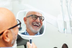 Patient with dental implants smiling into handheld mirror in dentist’s chair.